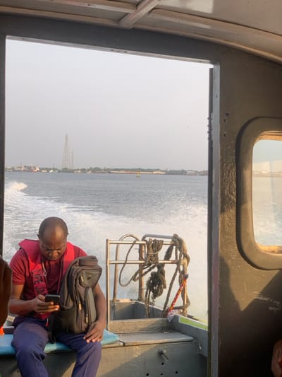 Inside a boat, a man in a red shirt and life jacket is engrossed in his phone. A black backpack rests on his lap. Outside the boat's window, water churns as the boat speeds along. The coastline is visible in the distance, with buildings and structures dotting the horizon. A transmission tower stands out against the hazy sky. Ropes and metal railings are attached to the boat near the window.
