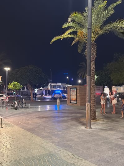 The image depicts a nighttime scene in Sant Antoni de Portmany, Spain, likely a promenade or street. The foreground is dominated by the textured paving stones of a sidewalk and a large palm tree. Several people are visible, some walking and others standing.

In the mid-ground, there's a kiosk or small building with advertising posters displayed. Further back, a street or road is visible with parked vehicles, including what appears to be a police car with blue flashing lights. Trees and shrubbery are scattered throughout the scene, providing a natural element against the urban environment. Bicycles are parked in racks, suggesting this is an area frequented by pedestrians and cyclists.

The lighting indicates it is nighttime, with artificial lights illuminating the area, casting reflections on the wet pavement. The overall mood appears relaxed and casual, consistent with a public space in the evening. The visible text on a poster is too blurry to decipher. The presence of palm trees and the general atmosphere suggest a Mediterranean coastal town.