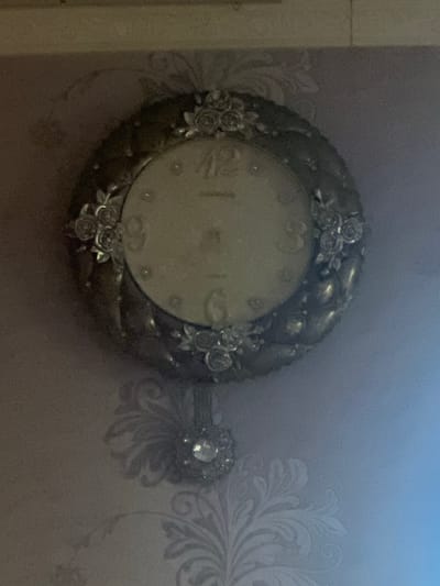 The image features a close-up of a decorative wall clock. The clock has an ornate, antique-style metallic frame adorned with sculpted roses and leaves. The clock face is a light, creamy color with Roman numerals, though only a few are clearly discernible, such as 'II' and 'III'. The hands of the clock are not visible. To the right of the clock, there is a decorative element attached to the wall, possibly a wall sconce or another decorative piece, featuring a large, faceted gem. The background is a wall with a subtle, damask-like floral wallpaper pattern in a lighter shade, suggesting an indoor setting. The lighting is dim, with a gradient of light from left to right, creating a slightly moody atmosphere. There is no visible text in the image. The scene is static, with no activity taking place. The focus is entirely on the clock and its immediate surroundings. The location context of Bujum, Kyrgyzstan, implies this could be found in a home or a public space within that region.