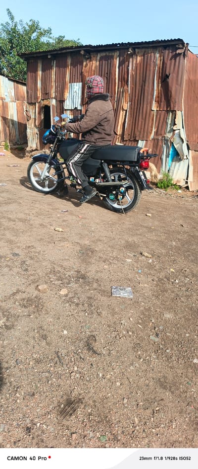 In Barikin Gidar Kanat, Nigeria, a person wearing a brown jacket, black pants with white stripes, and a plaid hat is seated on a dark-colored motorcycle. The motorcycle has silver wheels and chrome accents. Behind the motorcycle, a corrugated metal structure, likely a dwelling or workshop, shows signs of rust and damage. The ground is composed of dirt and small stones, with scattered debris including a piece of corn and a plastic wrapper. The sky is clear and blue. The scene appears to be in an outdoor, possibly rural or semi-urban, environment.