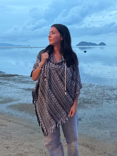 A young woman stands on the shore in Baan Sri Thanu, Thailand. She is wearing grey jeans and a patterned black and white poncho-style top with fringe. She has long dark hair and a black headband. She looks down and to the left. The ocean stretches out behind her, and in the distance, the silhouettes of mountains are visible under a cloudy sky. The ground is sandy.