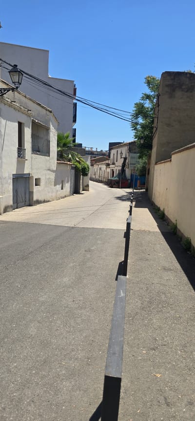 A vertical, eye-level, medium shot captures a street scene in Huesca, Spain, under a clear blue sky. The street is asphalt, with a lighter, more textured section bordering the right side, separated by a continuous dark metal railing. On the left, a white building with a rough, textured facade features a closed gray door and a window with a metal grate. Above it, a black lantern hangs from a decorative bracket. Further down the street, other white buildings with tiled roofs line the road. A lush green plant grows against a wall on the left. On the right, a cream-colored building wall casts a long shadow across the lighter pavement. The road curves slightly to the left in the distance, where more buildings are visible, along with trees and what appears to be utility poles and wires. The overall impression is a sunny, quiet street in a Spanish town.