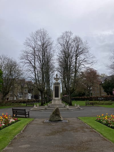 This media file captures a serene view of a public park or garden, likely in Bradford, United Kingdom, under an overcast sky. The image is composed with a central war memorial, flanked by tall, bare deciduous trees, suggesting late winter or early spring.
The war memorial stands prominently in the middle distance, featuring a substantial stone base with dark, rectangular plaques that likely bear inscriptions, though the text is unreadable from this distance. An ornate finial or urn crowns the memorial. In front of it, a large, natural stone boulder rests on the ground.
Asphalt paths wind through the park. One path curves to the right, bordered by neatly manicured green grass. Along this border, vibrant flower beds display a mix of colorful blooms, including red, yellow, pink, and white flowers. Another path is visible to the left, where several dark wooden park benches are placed, offering seating for visitors. Small white flowers, possibly daisies, dot the grassy areas.
In the background, behind the trees and the memorial, several multi-story stone or brick buildings with numerous windows are visible, characteristic of British architecture. The sky is uniformly gray and cloudy, indicating an overcast day. No people or activities are visible, contributing to the tranquil and somewhat somber atmosphere of the scene.