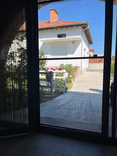 The image shows an outdoor scene viewed through an open window or doorway. Outside, there is a white building with a red tile roof under a clear blue sky. A concrete path leads from the building to a small white wooden fence, behind which are plants with red and pink flowers. To the right, there is another building or structure partially visible. The interior space, where the photo is taken from, is dark with horizontal blinds partially obscuring the view below the window frame. The scene is likely in Blatska, Bulgaria, as indicated in the prompt.