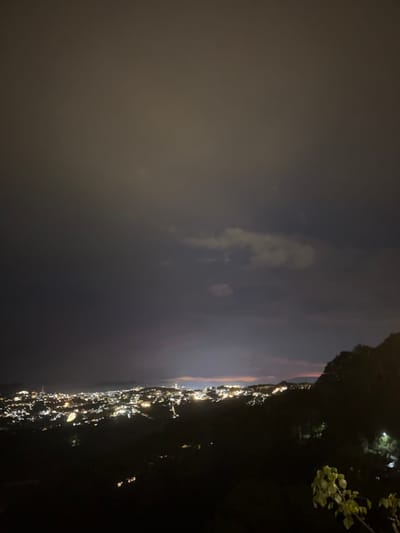 This is a night-time, high-angle, wide shot of a coastal town at night. The scene is dominated by the glow of numerous lights from the town, stretching along a coastline. The lights are mostly white and yellow, creating a luminous band against the dark landscape and the night sky.
The sky is dark with some wispy clouds illuminated by the town's lights, and a faint reddish glow is visible on the horizon, suggesting either a recent sunset or artificial lighting. To the right of the frame, a dark, silhouetted hillside covered in vegetation is visible, with a few scattered lights higher up on its slopes. A single branch with light green leaves is also visible in the upper right portion of the image, silhouetted against the sky.
The image appears to have been taken from a vantage point overlooking the town, possibly from a hill or a tall building. The overall mood is serene and tranquil, capturing the quiet beauty of a town at rest under the night sky. No people or specific objects are clearly identifiable due to the low light conditions and the distance. There is no visible text in the image. The location context is Xuân Thành, Vietnam, which suggests a Southeast Asian coastal setting.