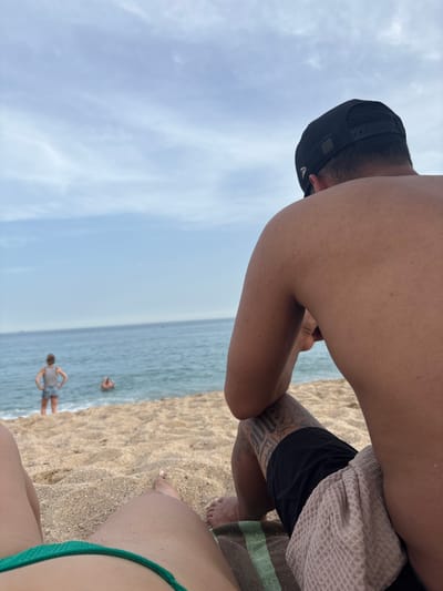 The image depicts a scene at a beach, likely in Vilassar de Mar, Spain. In the foreground, the lower body of a person wearing a green bikini bottom is visible, lying on the sand. To the right of this person, the feet of another individual are visible, resting on a striped towel and a waffle-weave towel. Dominating the right side of the image is a person from the back, wearing a black baseball cap and black swim trunks, with tattoos visible on their leg. A light-colored waffle-weave towel is draped over their leg. In the mid-ground, the ocean stretches out, with gentle waves lapping the shore. Two other individuals are in the water; one, a woman wearing a striped top and denim shorts, stands near the water's edge with her hands on her hips, while another person is partially submerged in the water further out. The background consists of a hazy sky with scattered clouds. The beach itself is sandy, with some small pebbles and debris. The overall impression is a relaxed day at the beach.