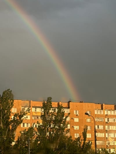 A rainbow arcs across a dark, overcast sky above a multi-story brick apartment building in Volzhsky, Russia. The building's facade is a warm orange-brown, punctuated by numerous windows of varying sizes, some with air conditioning units attached. Green trees with dense foliage are visible in the foreground, partially obscuring the lower floors of the building. A street lamp stands to the right of the building, its dark pole extending upwards towards the sky. The overall impression is a serene post-rain scene in an urban environment.