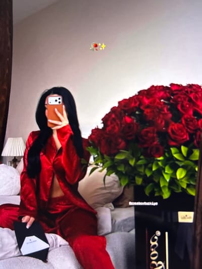 The image depicts a person in red satin pajamas sitting on a bed, holding a phone in their right hand to take a mirror selfie. To the right of the person is a large bouquet of red roses in a black box. The box has some text on it, possibly a company name or logo. The room appears to be a bedroom with a lamp visible on the left side of the frame. The overall impression is one of luxury and romance. The provided location information "Beșalma, Moldova" suggests this scene might be taking place in a private residence in that region.