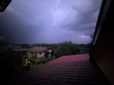 The image captures a nighttime lightning storm over Zornitsa, Bulgaria, viewed from an elevated indoor position. The immediate foreground is dominated by dark reddish-brown roof tiles, with a segment of a brighter pink-red roof visible to the right. In the midground, a light-colored house with a red-tiled roof and dark window frames is prominent on the left, nestled among dark foliage with a subtle point of artificial light. Another red-roofed structure is partially obscured behind it. The upper half of the frame displays a dark, purplish-gray, heavily overcast sky, indicative of storm conditions. Multiple distinct white lightning strikes are visible on the right side, illuminating sections of the clouds. A faint, distant landscape with minimal scattered lights is discernible on the horizon. Dark architectural structures, likely eaves or a window frame, partially obstruct the top-left and top-right corners.