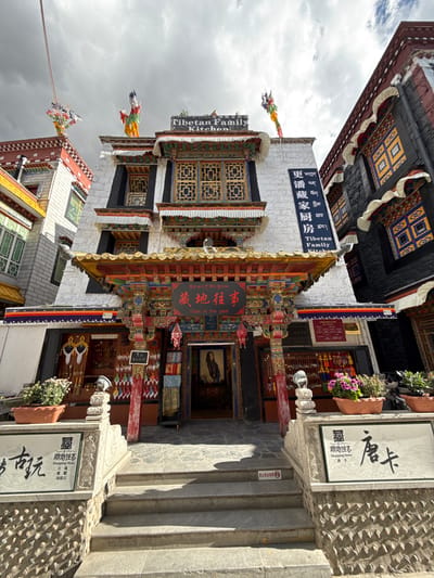This image captures a vibrant street scene in Chengguan District, featuring buildings with distinct Tibetan architecture under an overcast sky. The setting appears to be a commercial area, likely a street lined with shops and eateries.
The buildings showcase intricate details, including colorful painted wooden eaves with geometric patterns and ornate window frames. The facades are primarily white stucco or stone. Numerous prayer flags, in traditional colors, are strung across the street and affixed to the buildings, adding to the cultural atmosphere.
Several signs are visible. A prominent white sign on a building reads "Tibetan Family Kitchen" in English and "更潘藏家厨房" (Gengpan Tibetan Family Kitchen) in Chinese. Another building features a sign with "藏地往事" (Tibet in the past) in Chinese and English, marking a shop. Below it, a white rectangular sign further identifies it as "藏地往事 Shopping Store," listing "天珠 蜜蜡 绿松石" (Dzi beads, beeswax, turquoise) and "唐卡" (Thangka) as items sold. Flower pots with purple blooms adorn the entrances, adding a touch of natural beauty.
The ground features stone paving, with some areas having embedded pebbles, and a set of stone steps leads up to an entrance on the right. Small red signs with a white symbol and Chinese characters "禁止商业拍照" (No commercial photography) are visible, indicating restrictions on commercial picture-taking. No people are explicitly visible, suggesting the focus is on the architecture and streetscape. The diffuse light indicates daytime under cloudy conditions.