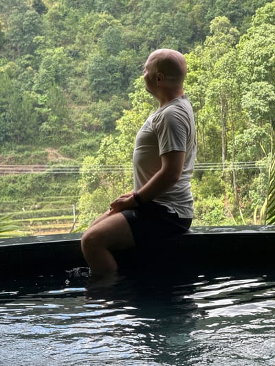 A bald man, viewed from the side, sits on a dark stone ledge with his lower legs and feet submerged in rippling water, facing left towards a verdant mountainous landscape. He wears a light grey short-sleeved t-shirt and dark shorts, with a multi-colored bracelet on his left wrist. The water reflects shimmering light, indicating an outdoor setting, likely an infinity pool or natural spring. In the background, the scene reveals lush green, densely forested mountains, characteristic of the Mù Cang Chải region in Vietnam. Several tiers of agricultural terraces, consistent with rice paddies, are carved into the lower slopes of the mountains. Three parallel utility lines are visible horizontally across the midground of the landscape. The overall impression is one of a man relaxing and contemplating the natural, agricultural scenery.