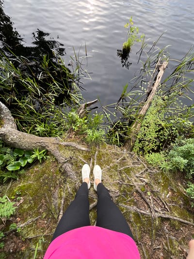 The image shows a person's legs and feet wearing white sneakers and black leggings, standing on mossy ground with exposed tree roots near the edge of a body of dark water. The water has some vegetation growing in it and along its edge, with a piece of driftwood visible. The scene appears to be a natural outdoor environment. The location is identified as the city of Nagorny, Russia.