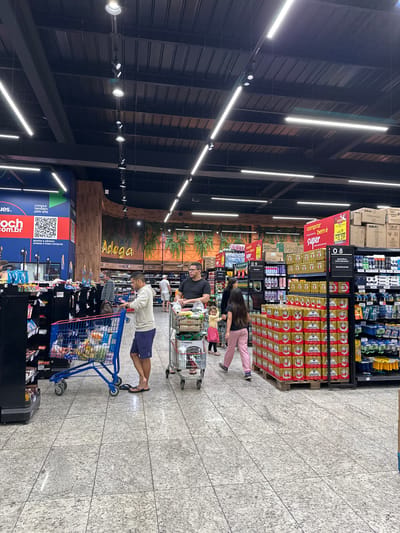 The image depicts the interior of a grocery store, specifically focusing on a section with beer and other goods. Overhead are linear lights, and the store features a dark, modern design. Several customers are present, each pushing a shopping cart filled with groceries. The shelves are stocked with various products, including beer cans arranged in stacks, deodorants, shaving products, and other miscellaneous items. A large sign above the shelves reads "Adega," and another display with a QR code invites customers to shop online. The floor has a speckled pattern. The image was taken at Koch supermarket in Florianópolis, Brazil.