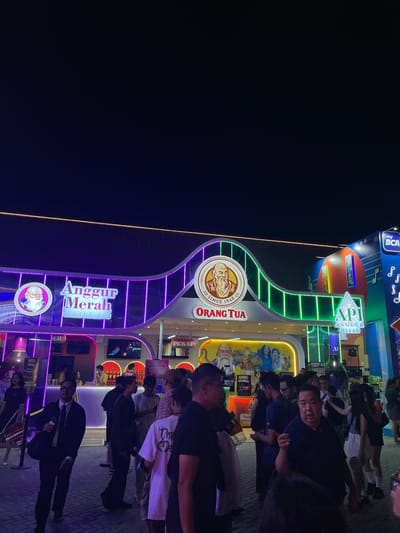 The image shows an outdoor nighttime scene in Salembaran, Indonesia, likely at a public event or fair. A large, brightly lit archway serves as a focal point, adorned with the "Orang Tua" and "Anggur Merah" logos. The archway also displays neon lights, and the surrounding structures have similar colorful lighting. A crowd of people is gathered near the structure. Some of them are looking at the camera. The ground is paved with light-colored bricks or tiles. The background is mostly dark.