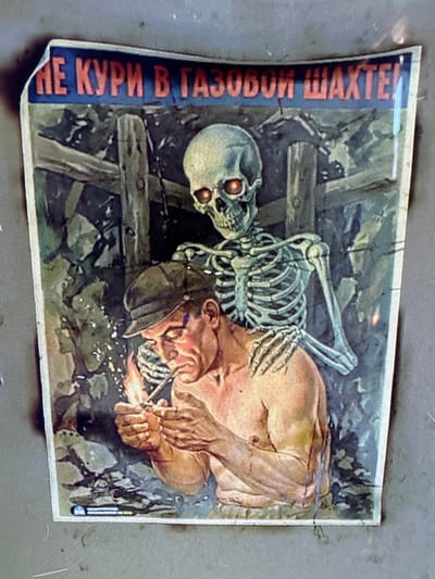 This image depicts an old, vertically oriented safety or propaganda poster, likely from a Russian-speaking region, warning against smoking in mines. The central subject is a muscular man, possibly a miner, who is in the act of lighting a cigarette with a match. Sparks and a small flame are visible where he strikes the match. He wears a cap and appears shirtless or partially clothed.

Looming beside and slightly behind the man is a skeleton, serving as a chilling representation of death. The skeleton's eye sockets glow with an ominous orange-red light, directing its gaze towards the man. Its bony hand is positioned close to the man's arm, suggesting an impending threat or a direct warning.

The scene is set in a dark, confined space, characteristic of a mine shaft, with rough-hewn wooden beams and supports visible in the background, reinforcing the underground setting. The ground beneath the figures is rugged and uneven. The overall atmosphere is dark and cautionary.

A prominent blue vertical strip on the left side of the poster contains the warning text in red Cyrillic letters. The text reads: "НЕ КУРИ В ГАЗОВОЙ ШАХТЕ!", which translates to "DO NOT SMOKE IN A GAS MINE!". The poster's message is a stark admonition against the dangerous act of smoking in potentially explosive environments, using the vivid imagery of death to underscore the fatal consequences. The image appears aged, with faded colors and slightly worn edges.