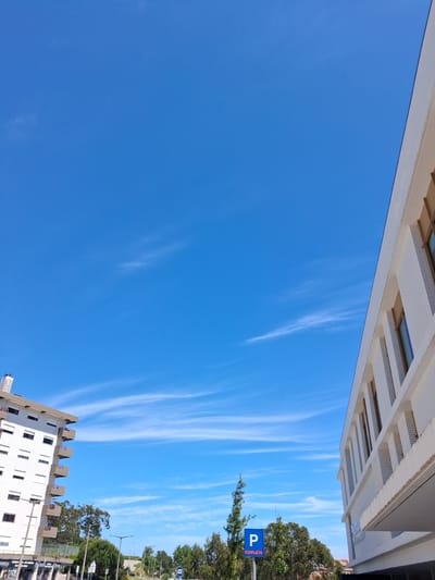 The image depicts a bright blue sky with wispy cirrus clouds, framed by buildings and greenery.
In the upper portion of the image, a building with a white facade and distinct rectangular design elements is visible. On the right side of the image, trees and vegetation line the area. A blue parking sign with the word "COMPLETO" is visible among the trees.
In the lower portion of the image, a tall white building with multiple windows stands.
The image was taken in Figueira da Foz, Portugal.