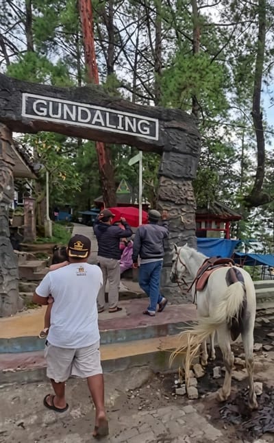 The image shows an entrance archway with "GUNDALING" written on it, indicating the location is Gundaling 2, Indonesia. A white horse with a saddle is standing to the right, facing away from the camera. Several people are present: a man in a white t-shirt and shorts is carrying a child, walking away from the camera. Two other men are standing near the archway, one holding a red umbrella. In the background, there are trees, a red-roofed building, and some blue tents or awnings. The ground is a mix of dirt and paved surfaces with some scattered stones.