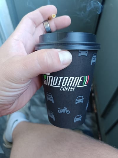 A person's hand, adorned with a silver ring on the index finger, holds a black paper coffee cup. The cup features a repeating pattern of stylized vehicles, including cars, a jeep, and a motorcycle, in a light blue color. The prominent logo on the cup reads "il MOTORE COFFEE," with the Italian flag colors incorporated into the design. A lit cigarette is held between the thumb and index finger, with smoke gently rising upwards. The background appears to be an outdoor setting, possibly near a building, with muted colors. In the lower left portion of the image, a white sock and part of a shoe are visible, suggesting the person is wearing casual attire. The scene is captured from a close-up perspective, focusing on the hand, cup, and cigarette. There is no information in the image to suggest it is located in Arkhangelsk, Russia.