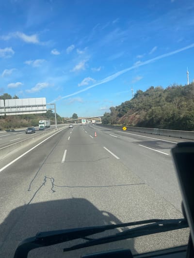 The image depicts a multi-lane highway during daytime, observed from the interior of a vehicle, likely near Sant Julià de Ramis, Spain. The foreground shows the vehicle's dashboard and windshield wipers, with a distinct dark shadow of the vehicle cast on the asphalt road surface. Irregular black tar lines are also visible on the pavement.

The highway consists of at least three lanes of travel in the depicted direction, marked by white dashed and solid lines. A single orange and white traffic cone is centrally positioned in the middle lane. A yellow circular sign, indistinct in detail, is visible on the right shoulder behind a concrete barrier that separates traffic lanes. Additional vehicles are present on the highway: a dark-colored sedan and a white truck with a green container are in the leftmost lane in the midground, with other vehicles in the distance.

In the background, a large overhead gantry structure with multiple sign panels spans the highway on the left. A concrete overpass crosses the highway further ahead. Hilly terrain, densely covered with green and reddish-brown vegetation, flanks both sides of the road. Several communication towers are discernible on the hill to the right. The sky is bright blue with scattered white clouds and a prominent white contrail extending diagonally across the upper frame. No individuals are visible in the scene.