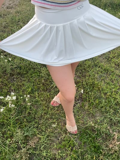 A low-angle shot shows a person's legs and white skirt outdoors in a grassy area. The skirt is pleated and flares out, obscuring most of the person's body. The person is wearing light-colored sandals with a buckle detail and has painted toenails. The background consists of green grass and some small white wildflowers. No specific location in Coșnița, Moldova, is identifiable from the image.