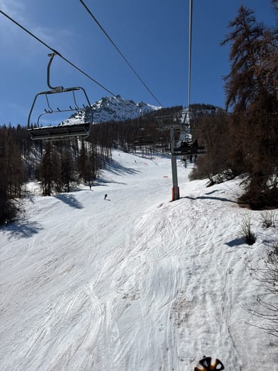 This image depicts a snow-covered ski slope and lift infrastructure on a clear, sunny day in Saint-Chaffrey, France. The perspective is from an elevated ski lift chair, with a portion of a black and yellow ski helmet visible in the bottom right corner.

In the mid-ground, a prominent empty grey metal ski lift chair hangs from taut cables stretching across the upper frame. Below, the wide ski slope descends, showing extensive ski tracks and some visible dirt patches on the snow. Several dark-clad skiers are observable on the slope: one actively descends left of center, another is further up-slope to the left, and a third small figure is visible higher up. A grey ski lift support pole with a red base stands on the right side of the slope, supporting the cables. Beyond this pole, another ski lift chair carries at least two occupants.

The slope is flanked by dense forests featuring mostly bare deciduous trees and some darker coniferous evergreens. In the background, rugged, snow-capped mountain peaks rise against a vibrant, cloudless blue sky. Strong sunlight casts distinct shadows from trees and ski lift components onto the snow.