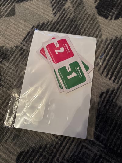 The image shows a set of screen protectors on a gray carpet. The screen protectors are white and rectangular, and they are still in their plastic packaging. There are also two small packets of wipes, one red and one green, which are also part of the screen protector kit. The red packet is labeled "Dry" and has a large number "2" on it. The green packet is labeled "Wet" and has a large number "1" on it. Both packets are labeled "Professional Screen Cleaning Paper" and "wipes removes excess dirt". The image is taken from a high angle, looking down at the screen protectors. The lighting is natural, and the focus is on the screen protectors. There is no indication of the location in Chernogorsk, Russia.