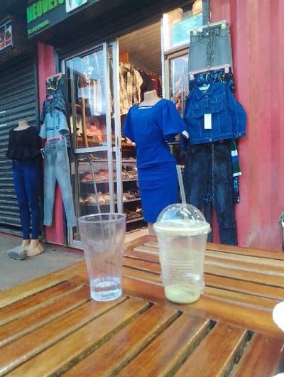 The image shows a wooden table with two empty glasses and a plastic cup with a green liquid, situated outside a clothing shop in Rungiri, Kenya (coordinates: -1.25, 36.684). The shop front is visible, displaying a variety of clothing items, including jeans, shirts, and a blue dress on a mannequin. The shop's facade features a red metal wall with a sign in the upper left corner. The floor is made of cement tiles.  There are two mannequins outside the shop, both female, dressed in jeans and shirts. One of the mannequins is wearing a blue dress. The scene suggests a retail setting with a focus on clothing, where the table is likely used for customer seating or product display.  The image captures a moment of quiet business activity, with no visible patrons.  The light source appears to be coming from the shop windows, creating a slightly dim atmosphere.