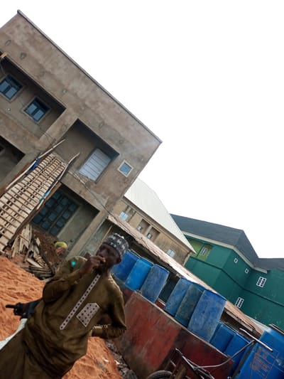 The image depicts an outdoor scene in Arawa, Nigeria, dominated by construction activities and storage of large blue barrels. In the foreground, a man wearing a brown, patterned garment and a black and white cap is partially visible, appearing to hold something to his mouth. He is standing on sandy ground. Behind him, a partially constructed concrete building with several windows is visible. To the right, a row of large blue plastic barrels is stacked against a red wall. Further in the background, another multi-story building, painted green, stands with its roof visible. The overall environment suggests a construction site or storage area in an urban setting.