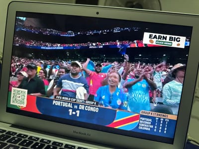 A laptop screen displays a live broadcast of a soccer match, with a score overlay indicating "PORTUGAL vs DR CONGO 1-1". The broadcast is for "FIFA WORLD CUP 2026 - GROUP K". In the foreground of the broadcast image, a crowd of enthusiastic spectators can be seen. One spectator in the front row is wearing a blue jersey. There is an advertisement banner at the top right of the screen reading "EARN BIG CLICK HERE!". A QR code is also visible on the left side of the screen. The broadcast also shows a "GROUP K STANDINGS" table. The laptop itself is open, with a keyboard visible at the bottom of the frame. The scene appears to be indoors, likely a room in Kufena, Nigeria, given the context.