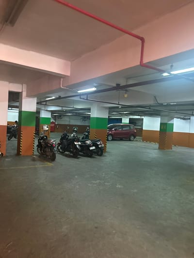 The image depicts an indoor parking garage, possibly in Makassar, Indonesia, indicated by a partial view of a red car with a license plate visible, though the text is illegible. The garage has a concrete floor and ceiling, with fluorescent lighting. Red pipes run along the ceiling, suggesting a fire suppression system. Several motorcycles are parked in designated spots, some with helmets on them. A red MPV is parked in the background. Pillars painted with orange, green, and yellow stripes mark the parking bays. There are no visible individuals engaged in any activity, and the scene is static. The overall atmosphere suggests a typical, functional parking facility.