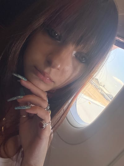 The image is a close-up, selfie-style photograph taken from inside an airplane, looking out the window. A young woman with long, dark brown hair and bangs is the primary subject. Her face is partially obscured by her hair and the angle of the shot. She is resting her chin on her hand, which features long, light blue, pointed fingernails decorated with gold flecks. She is wearing at least two rings on her fingers. The woman's eyes are visible and appear to be a light color, possibly green or grey. Her lips are slightly parted.
Through the airplane window, an outdoor scene is visible. There are buildings and structures that suggest an airport or a developed area. The sky is clear and bright, indicating daytime. The distinct curve of the airplane window is prominent, framing the outdoor view. The location is not specifically identifiable as Yagel, Israel, based on the visual information presented in the image alone.