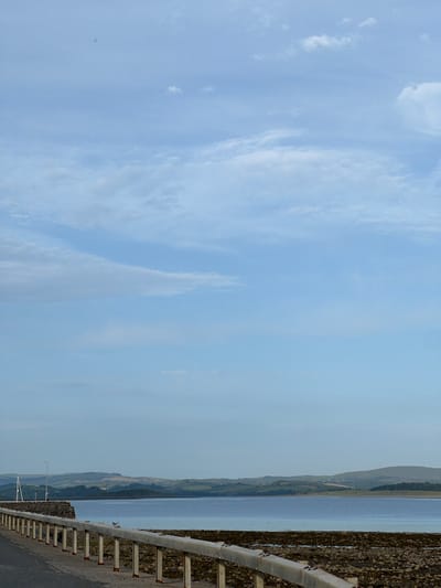 The image shows a view of a body of water with hills in the background. In the foreground, there is a paved surface with a metal railing. The water is calm and reflects the sky, which is partly cloudy. The hills are covered in vegetation and appear to be rolling. There is a stone structure on the left side of the image, and a lamppost visible behind it. The scene suggests a coastal or lakeside environment. No specific landmarks within Terhillion, Ireland are identifiable from this image.