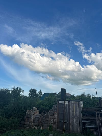 This media file captures a partly cloudy sky over a rustic scene in Tobyl, Kazakhstan. The main subjects are the sky and a collection of weathered wooden structures and materials.
The sky is a vibrant blue with distinct cloud formations. A large, billowy cloud stretches diagonally across the frame, casting shadows and highlighting its textured surface. Smaller, scattered white clouds dot the expanse. The overall impression of the sky suggests a bright day, likely late morning or early afternoon, with fair weather.
The ground level is dominated by an assortment of old, weathered wooden objects and building materials. These include what appear to be doors, planks, and possibly corrugated metal sheets, stacked and leaning against each other. Some of these items show peeling paint and signs of decay, indicating they are likely disused or part of a construction or demolition site. A pile of bricks is visible near the bottom right. Surrounding the structures are lush green trees and foliage, suggesting a rural or semi-rural setting. Power lines are also visible in the background, hinting at proximity to infrastructure.
There is no explicit activity or event taking place; the image appears to be a still-life composition of the environment. The scene evokes a sense of age and disuse, with the natural beauty of the sky contrasting with the man-made decay. The overall mood is calm and perhaps a little melancholic due to the state of the objects. No visible text is present in the image.