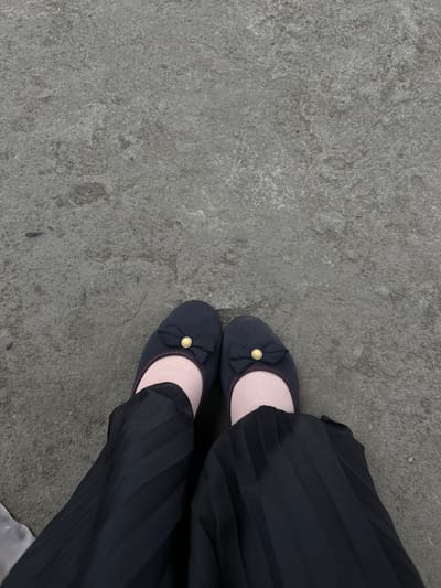 This is a close-up, top-down shot of a person's legs and feet, wearing dark blue ballet flats with a small bow and gold accent on the front, and black pleated pants. They are wearing pale pink socks. The ground appears to be a rough, grey concrete surface. The scene is devoid of any specific landmarks or contextual clues that would pinpoint its location in Palohlada, Indonesia.