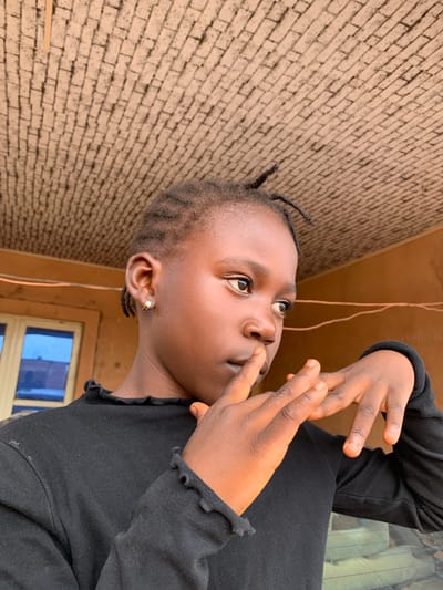 The media file shows a close-up, low-angle, portrait of a young Black girl. She is wearing a black long-sleeved shirt with ruffles at the collar and cuffs. Her hair is styled in thin braids, with a few braids pulled forward at her forehead. She has a small gold hoop earring in her left ear. Her face is turned upwards and to the right, and she is looking off-camera with a thoughtful or questioning expression. Her hands are raised to her face, with her fingers near her mouth, as if she is contemplating something.
The background consists of a brick wall, which appears to be textured and light brown in color, and a wooden surface above it. A thin, light-colored wire hangs down in front of the wooden surface. To the right, a portion of a window with blue panes is visible. The lighting suggests it is daytime, with light illuminating the girl's face from above. The overall setting appears to be outdoors or in a covered outdoor space, possibly a porch or patio, in Barakin Sabawa, Nigeria. The image has a slightly warm tone. There is no visible text in the media file.