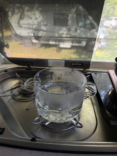 A glass mug filled with water is placed on a stovetop. The water appears to be heating up, with small bubbles visible on the surface and condensation on the glass. The stovetop is metallic and appears to be part of a mobile home or RV kitchen. In the background, a window is covered by a mesh screen, through which trees and vehicles are faintly visible. The scene suggests a domestic setting, possibly in Kazanlak, Bulgaria, given the context.
