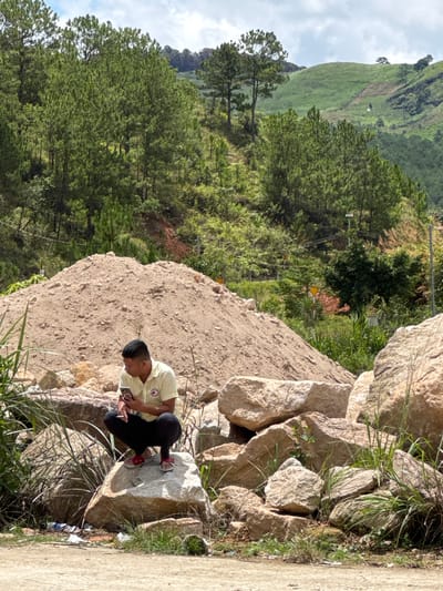 A man is squatting on a large rock, holding a mobile phone in his right hand. He is wearing a yellow short-sleeved collared shirt and black pants. The man is positioned in the foreground, with a large pile of sand behind him. In the background, there are steep, tree-covered hills. The environment appears to be rural or mountainous, with lush vegetation and a cloudy sky. There are some scattered rocks and grass around the man. The scene is consistent with the outskirts of Klong Klanh, Vietnam, a region known for its hilly terrain and agricultural or construction activities.