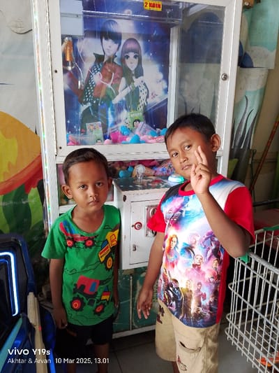 Two young boys are standing in front of a claw machine in a store in Kecamatan Haurgeulis, Indonesia. The claw machine has an image of two anime-style characters in the window. The boy on the left is wearing a green shirt with cartoon vehicles on it and has short, dark hair. He is looking down. The boy on the right is wearing a red and white shirt with cartoon characters on it and has short, dark hair. He is looking at the camera and has his right hand up near his face. The boys are both smiling. In the background, there is a shopping cart next to the claw machine. The store walls are covered in colorful designs. The image is taken with a cell phone camera. The time and date are displayed in the bottom left corner. The image is taken in the town of Kecamatan Haurgeulis in West Java, Indonesia, at coordinates -6.42 latitude and 107.955 longitude.