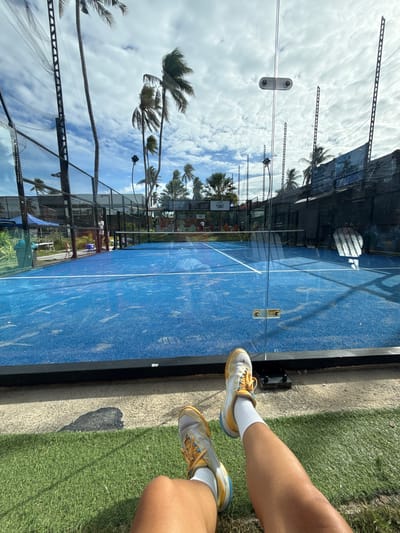 The image depicts a padel court located in Baan Plai Laem, Thailand. The court is vibrant blue with white boundary lines. A glass wall separates the court from a grassy area in the foreground where a person's legs and feet are visible, wearing white socks and yellow and white athletic shoes. Palm trees are scattered around the outdoor facility, and the sky is partly cloudy. The background shows fencing surrounding the court, buildings, and more palm trees, suggesting a tropical or subtropical environment.