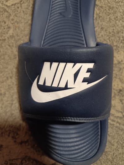 A close-up, top-down view of a navy blue Nike slide sandal is presented, lying on a carpeted surface. The sandal features the prominent white Nike swoosh logo and the word "NIKE" in white capital letters on the foot strap. The foot strap appears to be slightly worn or scuffed in places, with visible thin white lines suggesting minor damage. The sole of the sandal has a textured, ribbed pattern. The background is a neutral-colored carpet with a mottled texture. The image is taken indoors, and no specific location in Sucha Beskidzka, Poland, can be identified from the visual information provided.