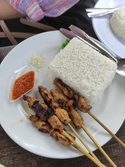 A white plate containing a meal of grilled chicken skewers, a mound of white rice, a small dish of chili sauce, and a sprinkle of sesame seeds. The skewers are on bamboo sticks and are arranged around the rice mound. The image is taken in a restaurant in Kecamatan Trawas, Indonesia, located at latitude -7.656 and longitude 112.594. The table surface is a dark brown wood. A portion of a striped shirt is visible at the top of the image. Two silver utensils are also visible in the background. The scene appears to be a meal ready to be eaten.