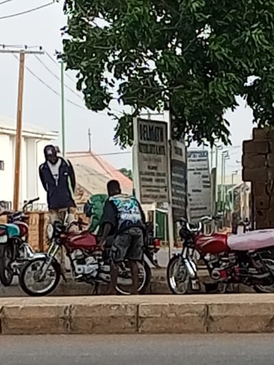 In Tumfure, Nigeria, two men are standing by motorcycles. One man, wearing a dark jacket with a white shirt underneath, stands behind a red motorcycle. The other man, in a dark shirt with a colorful design, is crouched down, possibly attending to a motorcycle. Several motorcycles are parked in a row along a curb, with some bearing luggage strapped to the back. A large tree with green leaves provides shade over the scene. Signs are visible in the background, suggesting a commercial area. The sky is overcast.