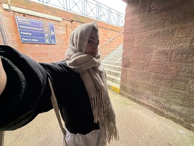 The image captures a young female standing in a pedestrian underpass or covered passage at a train station in Obermodern-Zutzendorf, France. The subject, positioned slightly left of center, is taking a selfie, holding the camera with her outstretched left arm, which is partially visible in the foreground. She wears a dark long-sleeved top, light-colored trousers, and a light beige/grey textured scarf covering her head and neck, with fringes visible on her left side. Her gaze is directed towards the camera with a neutral expression.

To the subject's left, a blue rectangular sign is affixed to a red brick wall. The sign displays white text and symbols indicating 'Sortie Exit • Ausgang', 'Correspondances', 'Parking', and 'Parc à vélos', along with directional arrows. The wall, constructed from reddish-brown, rough-hewn bricks, forms the left boundary of the scene. To the subject's right and extending into the background, a flight of concrete stairs ascends, bordered by a simple metal handrail. Above the stairs, a metal railing is discernible against an overcast sky. The ground beneath the subject is a light grey paved surface, likely concrete or asphalt, featuring a distinct yellow line at the base of the wall adjacent to the stairs.