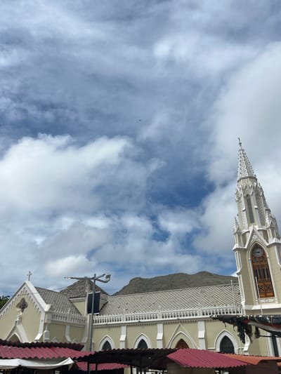 The image captures a portion of a light-colored church with ornate architectural details, including arched windows and decorative cornices. A prominent steeple, adorned with spires and what appears to be a clock face, rises towards a cloudy sky. The roof of the church is tiled with a repeating diamond pattern. In the foreground, a modern street lamp with two lights stands vertically. The scene is set against a backdrop of a rugged, gray mountain under a partly cloudy sky. The clouds are a mix of white puffs and thinner, grayish sheets, suggesting a daytime setting with variable weather. The orientation of the image is vertical, and the church appears to be viewed from a slightly elevated position. The location context provided is El Valle del Espíritu Santo, Venezuela. There is no visible text in the image.