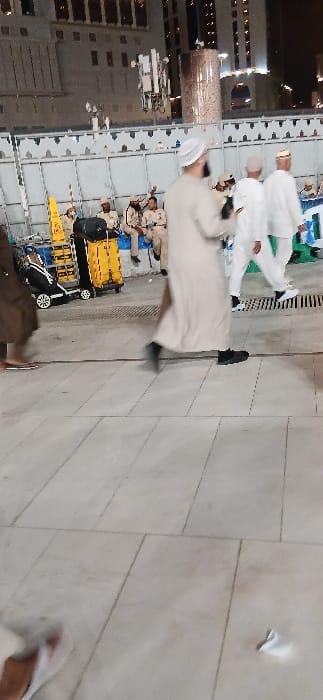 The image displays an outdoor pedestrian area in Makkah Al Mukarramah, Saudi Arabia, captured during the evening or night. The foreground consists of light-colored, large square-tiled pavement, with a crumpled piece of white paper visible on the lower right. Several male individuals are present. In the mid-ground, a man in a light beige thawb and white skullcap is walking from left to right, his motion blurred, with his left foot in a black shoe visible. Behind him, two other men in white thawbs and skullcaps are also walking. To the far left, a yellow cleaning cart, equipped with a "wet floor" sign and a black bag/container, is positioned. A light-colored barrier, featuring decorative upper cutouts, separates the foreground walkway from a group of approximately seven men in the background. These men are seated against the barrier, wearing light brown uniforms, dark trousers, and caps; some appear to be holding radios or phones. Overhead, against a dark sky, illuminated modern high-rise buildings are visible. A prominent slender pole supporting multiple communication antennas and a large cylindrical structure with a textured surface are situated in the mid-background.