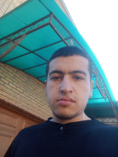An upward-angled close-up selfie shows a male individual, appearing in his late teens or twenties, centered in the frame. He has short dark hair, dark eyes, prominent eyebrows forming a unibrow, and light stubble on his chin and jawline. He wears a dark blue or black collared shirt with two visible silver buttons on the placket. His gaze is directed forward, meeting the camera.
The background behind the subject's left shoulder features a wall constructed of light beige or tan bricks. Below the brickwork, a horizontal decorative wooden panel with carved relief patterns is visible. A turquoise-colored translucent canopy or awning extends diagonally across the upper portion of the frame, supported by a dark metal frame with ornamental scrollwork visible on the right side. A bright, light-colored sky is visible above the canopy on the upper right. The scene is located in Kasan, Uzbekistan.