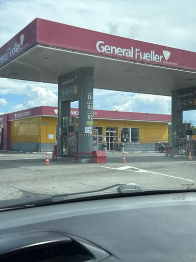 This is a medium-angle, outdoor, eye-level, full shot of a General Fueller gas station in Bolshiye Dvory, Russia. The gas station has a red roof and a yellow building with a red trim. The gas station has several pumps and a sign that says "General Fueller" and "REFEULLING SERVICE." There are orange traffic cones on the ground in front of the gas station. The sky is cloudy. The image appears to have been taken from inside a vehicle, as the dashboard and windshield wipers are visible in the foreground.
