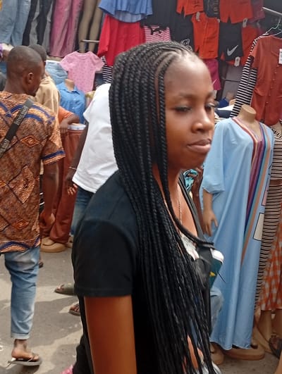 An outdoor market scene in Asaba, Nigeria, is depicted. The central figure is a young adult female, shown in a three-quarter profile facing right, with dark skin and long, thin black box braids. She wears a black short-sleeved t-shirt and has a white lanyard visible around her neck. Her gaze is slightly downcast with a neutral expression.

To the left and rear of the central figure, a male individual is partially visible from behind, wearing a brown and orange patterned short-sleeved shirt, blue jeans, and light-colored flip-flops. A dark strap crosses his left shoulder. Further in the background, another male figure wears a white t-shirt and blue jeans.

The background comprises a dense display of various garments, characteristic of a street market. Clothes hang on racks and are displayed on multiple mannequin torsos and full mannequins. Visible items include a light blue off-shoulder top, red blouses, dark t-shirts (one with a white Nike logo), a pink polka-dotted t-shirt, a light blue collared shirt, a light blue kaftan-style dress with vertical multi-colored stripes, and a patterned orange and white dress. The ground appears to be a paved surface. The scene is brightly lit.