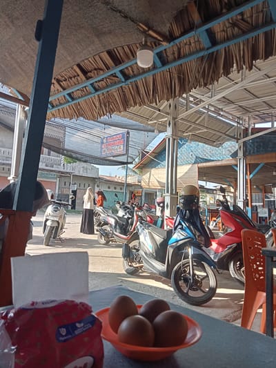 The image depicts an outdoor seating area of a food establishment in Meunasahalue Ie Puteh, Indonesia. The foreground features a table with a red package of "Kuliner" brand napkins and a small orange bowl containing three brown eggs. The middle ground shows several parked motorcycles, including a white scooter, a blue and black scooter, and a red scooter. Two women, one wearing a hijab and orange headscarf, and the other in a white hijab and dark skirt, are standing near the white scooter. In the background, buildings line a street, and a sign for "BAKSO CEKWAN BANDA" is visible. The establishment appears to have a thatched roof supported by blue metal beams and a black mesh awning overhead. The environment is sunny, with shadows cast on the ground.