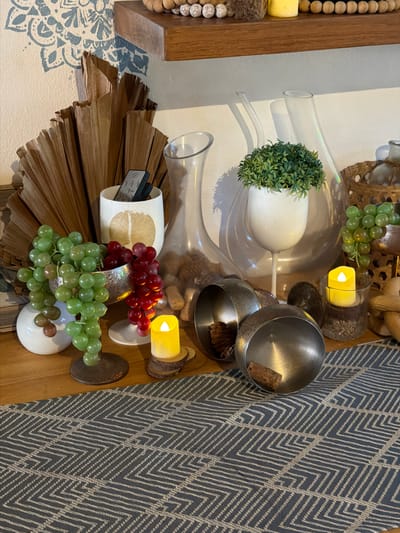 This is a close-up, slightly angled shot of a decorative arrangement on a wooden surface, likely a table or shelf, next to a patterned rug. The arrangement features several objects with a natural and rustic aesthetic.

Prominently displayed are bunches of artificial green and red grapes. Beneath the grapes are dried, fan-shaped palm leaves, creating a layered texture. A white cylindrical object holds a dark-colored remote control with visible buttons.

To the left, glass vases of varying shapes and sizes are stacked. One larger vase contains a small, artificial green plant. Further left, a wooden frame with wooden beads and a yellow cylindrical object is partially visible, suggesting this might be part of a larger piece of furniture or wall decor.

In the upper right, two metal bowls, one larger than the other, sit next to a cylindrical candle holder with a lit yellow candle. A piece of cork is visible in the larger metal bowl. The background includes a dark blue and beige patterned rug with a chevron design.

The lighting appears to be indoor artificial light, creating some shadows and reflections on the glass and metal objects. There are no people visible. The scene suggests a decorative display rather than an active event. The overall impression is one of curated interior design with natural and metallic elements. No legible text is present other than on the remote control. The location context is provided as Ko Tao Subdistrict, Thailand, which may inform the style of the decorative elements.